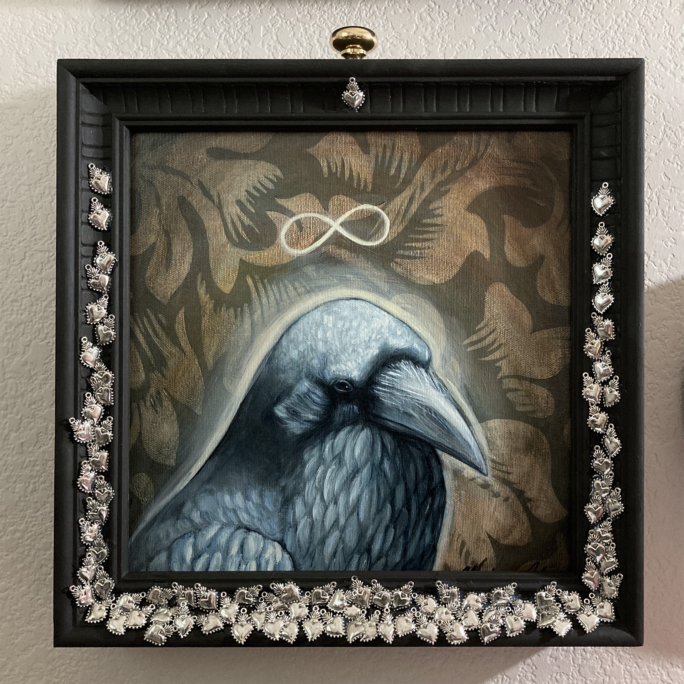 Painting portrait of a raven on a gray brown patterned background in a wood frame decorated with flaming heart charms.